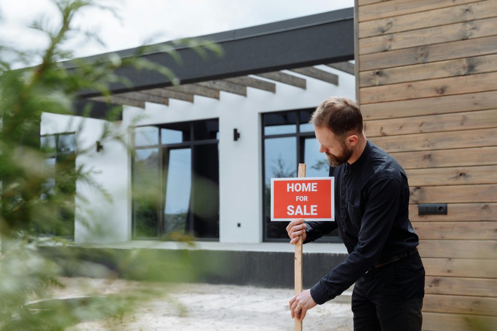 Man placing a home for sale sign outside a modern house for real estate listing.